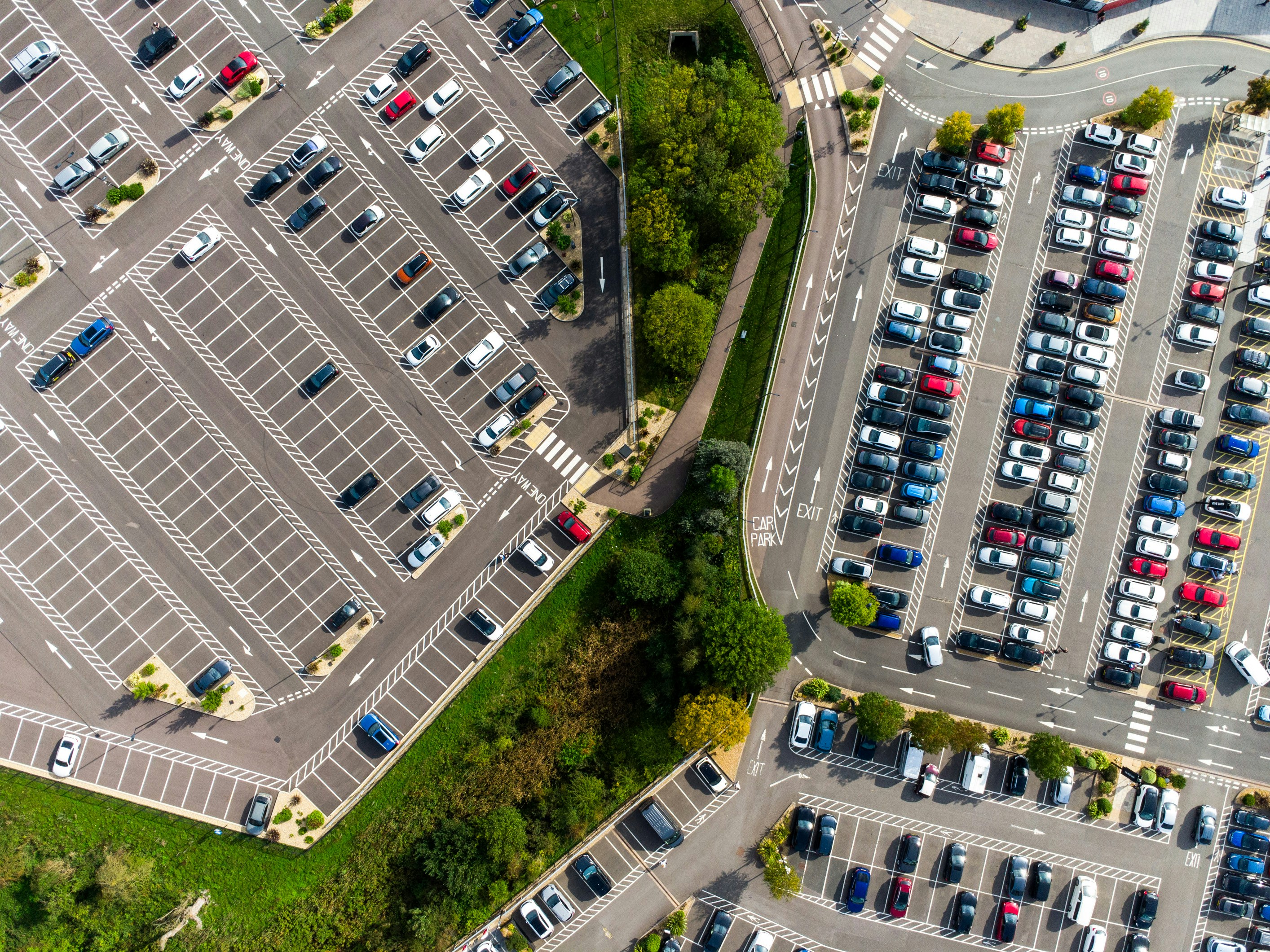 Aerial view of several busy car parks beside intersecting roads, showing rows of parked vehicles, marked bays, access routes and surrounding green spaces.
